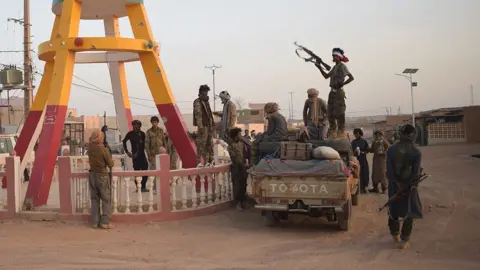 A group of armed fighters in camouflage stand around a military pick-up truck on a dusty road. One man near the vehicle’s edge is standing on the truck, holding an automatic rifle up. In the background on the left, the brown and yellow legs of a large sculpture are visible.