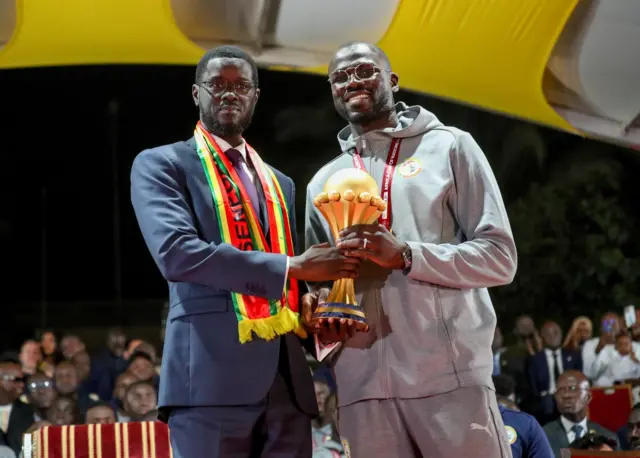 President Bassirou Diomaye Faye and captain Khalidou Koulibaly holding the CAN trophy together, wearing dark blue jackets and Senegal's national colors.